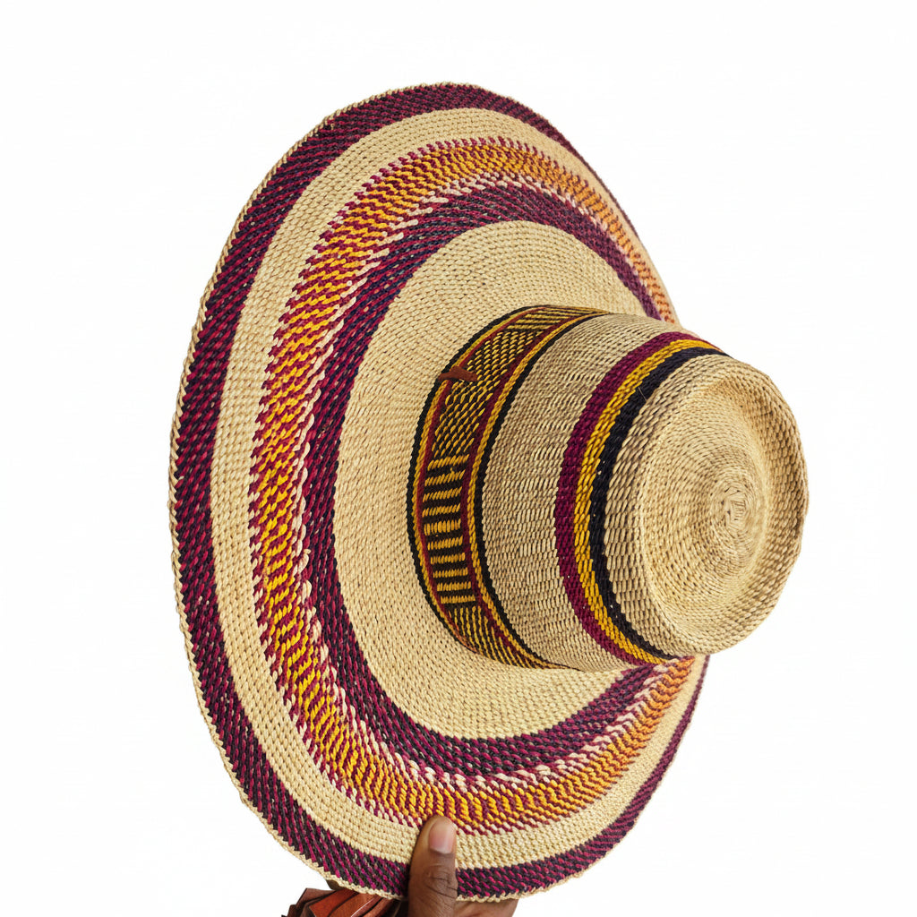 A woven straw boater-style hat with a wide brim, featuring concentric bands of purple, orange, yellow, and black woven patterns around a natural beige straw base. A hand is holding the brim on the right side in an indoor workshop or store setting.
