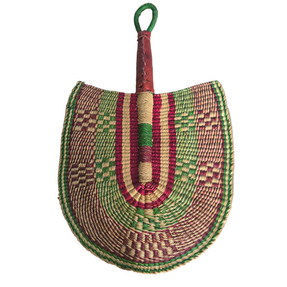 A hand-woven semicircular fan or shield-shaped accessory with a wrapped handle, placed on a white plastic tray. The woven body features concentric bands of green, red, beige, and brown in geometric patterns, with a green loop handle.