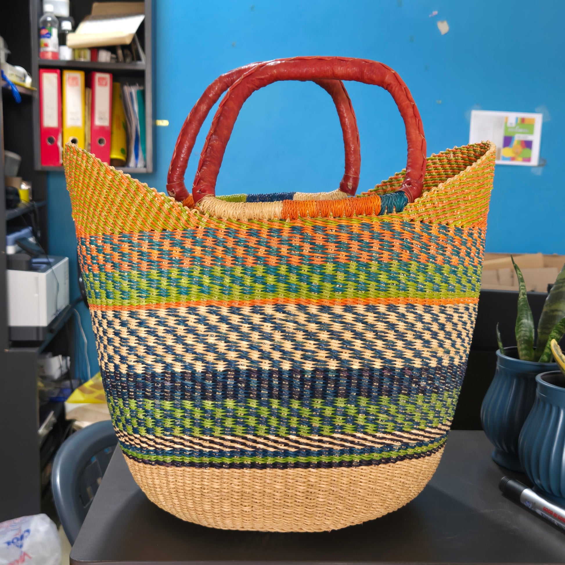 A woven basket bag with a natural tan base, multicolored striped patterns in green, blue, yellow, and orange, and reddish-brown double handles, photographed in an office-like setting.