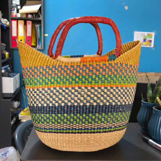 A woven basket bag with a natural tan base, multicolored striped patterns in green, blue, yellow, and orange, and reddish-brown double handles, photographed in an office-like setting.