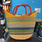 A woven basket bag with a natural tan base, multicolored striped patterns in green, blue, yellow, and orange, and reddish-brown double handles, photographed in an office-like setting.