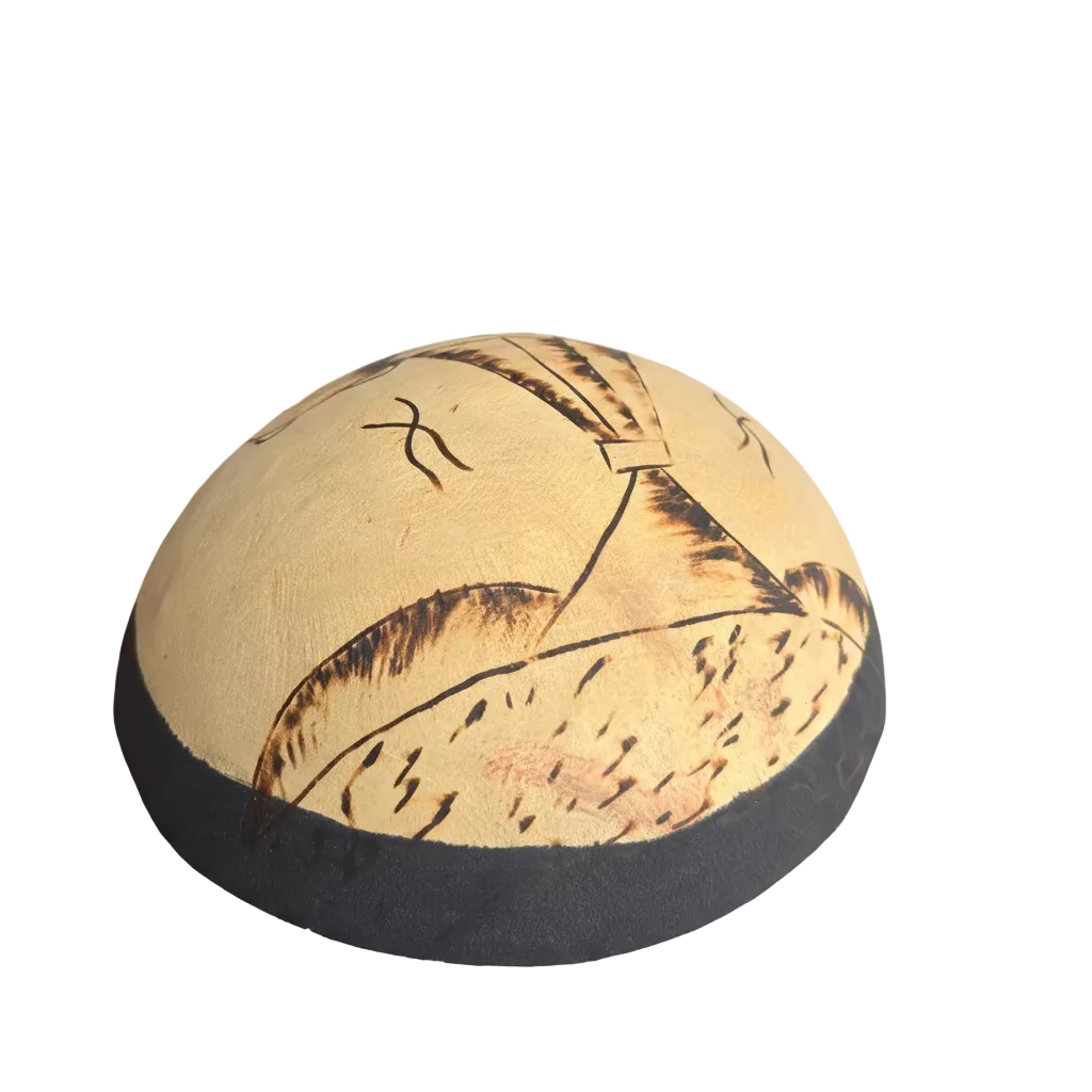 A hand holds a handmade round hat or cap made from natural beige palm leaves with dark brown burned or painted line details and a dark gray fabric trim.