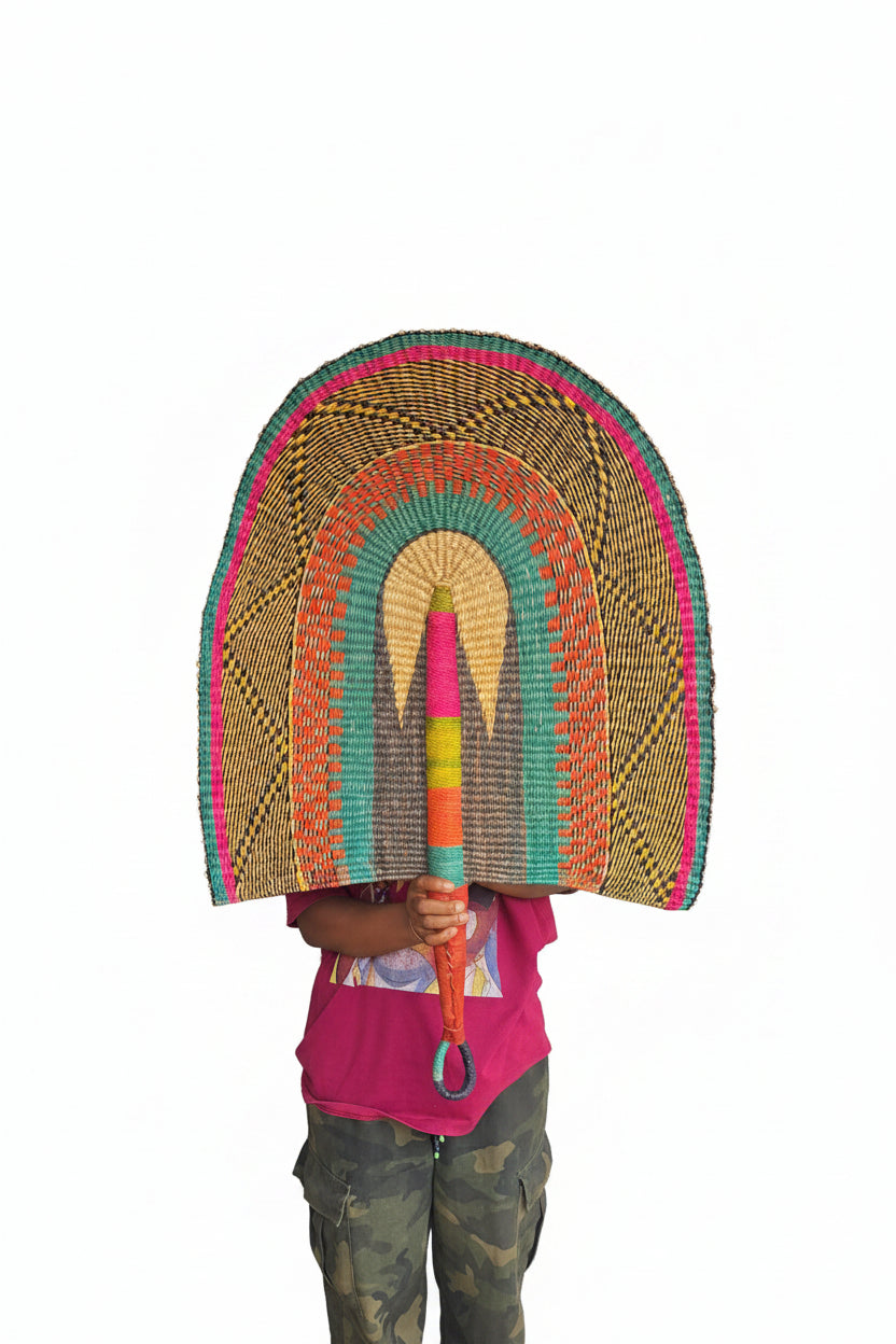 A person holding a large hand-woven straw fan with colorful concentric bands of pink, green, blue, and yellow, standing in a doorway.