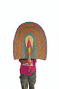 A person holding a large hand-woven straw fan with colorful concentric bands of pink, green, blue, and yellow, standing in a doorway.